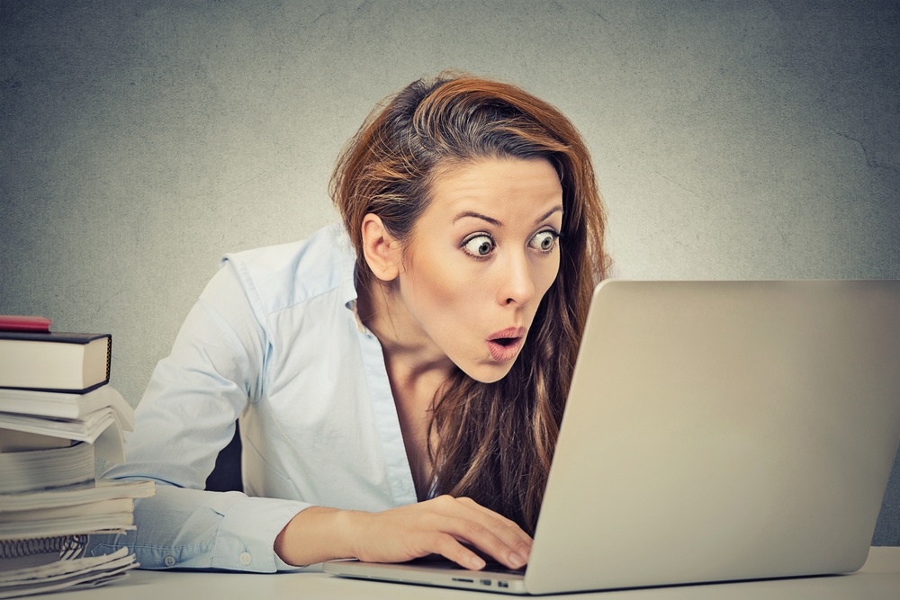 Portrait young shocked business woman sitting in front of laptop computer looking at screen isolated grey wall background. Funny face expression emotion feelings problem perception reaction .jpeg